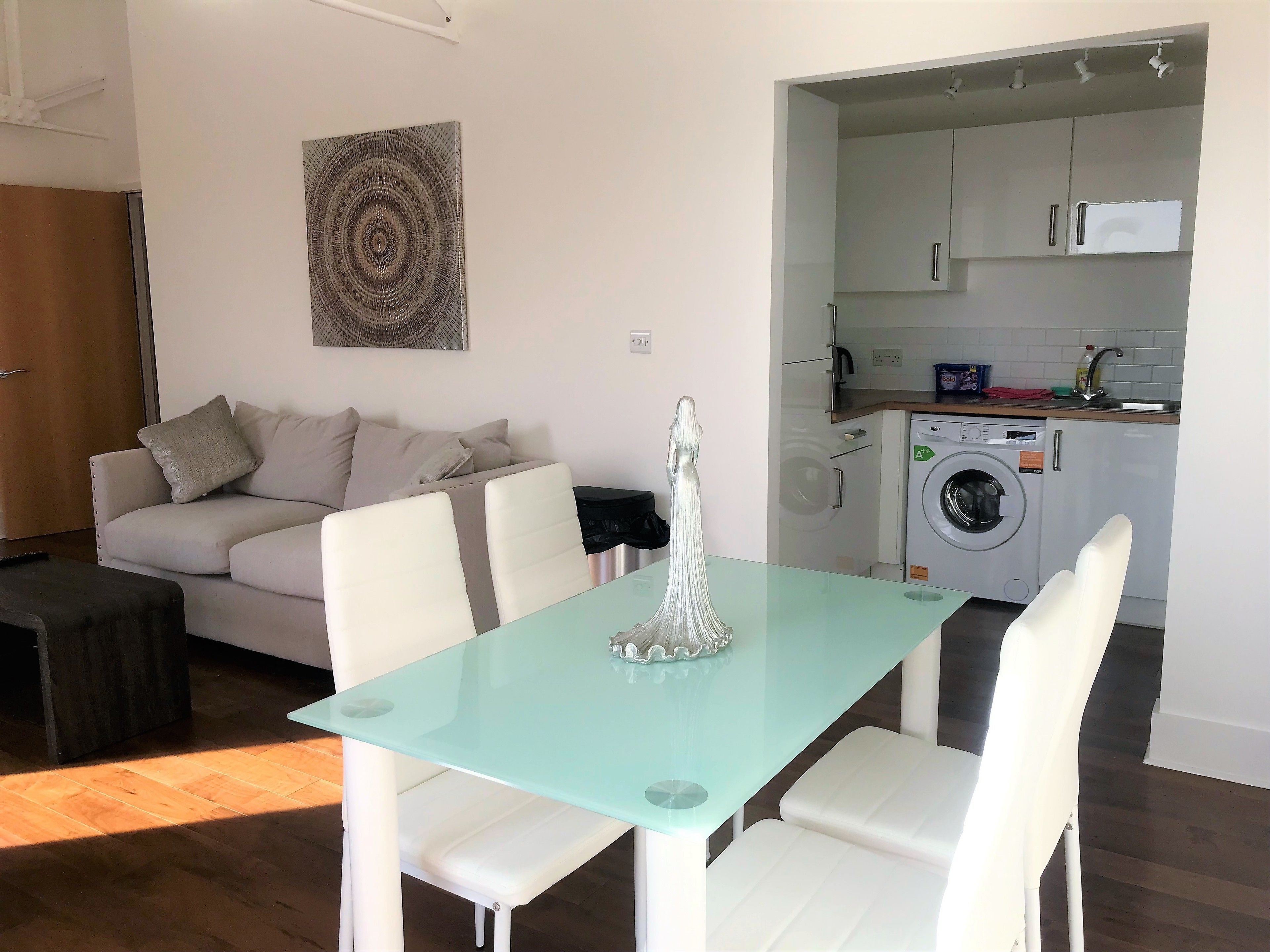 Una sala moderna con una mesa de vidrio, sillas blancas, un sofá y una lavadora al fondo.
