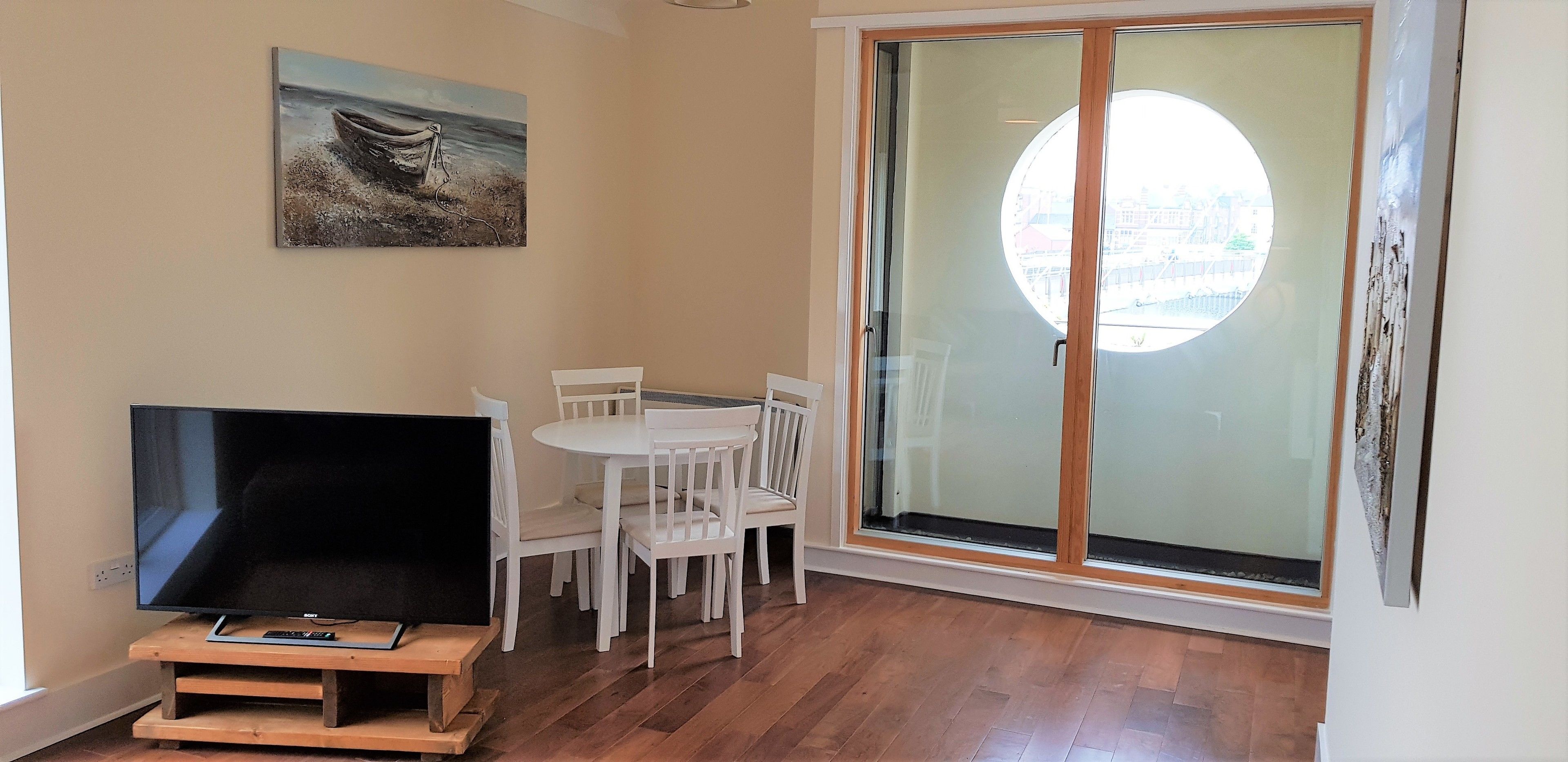 A room with a TV on a wooden stand, white chairs around a table, a painting on the wall, and a large glass door with a view.