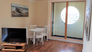 Een kamer met een televisie op een houten standaard, witte stoelen rond een tafel, een schilderij aan de muur en een grote glazen deur met uitzicht.