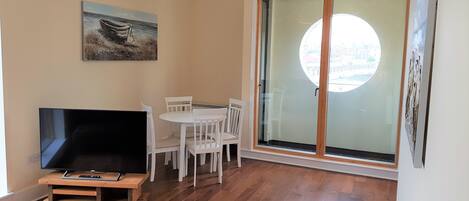 A room with a TV on a wooden stand, white chairs around a table, a painting on the wall, and a large glass door with a view.