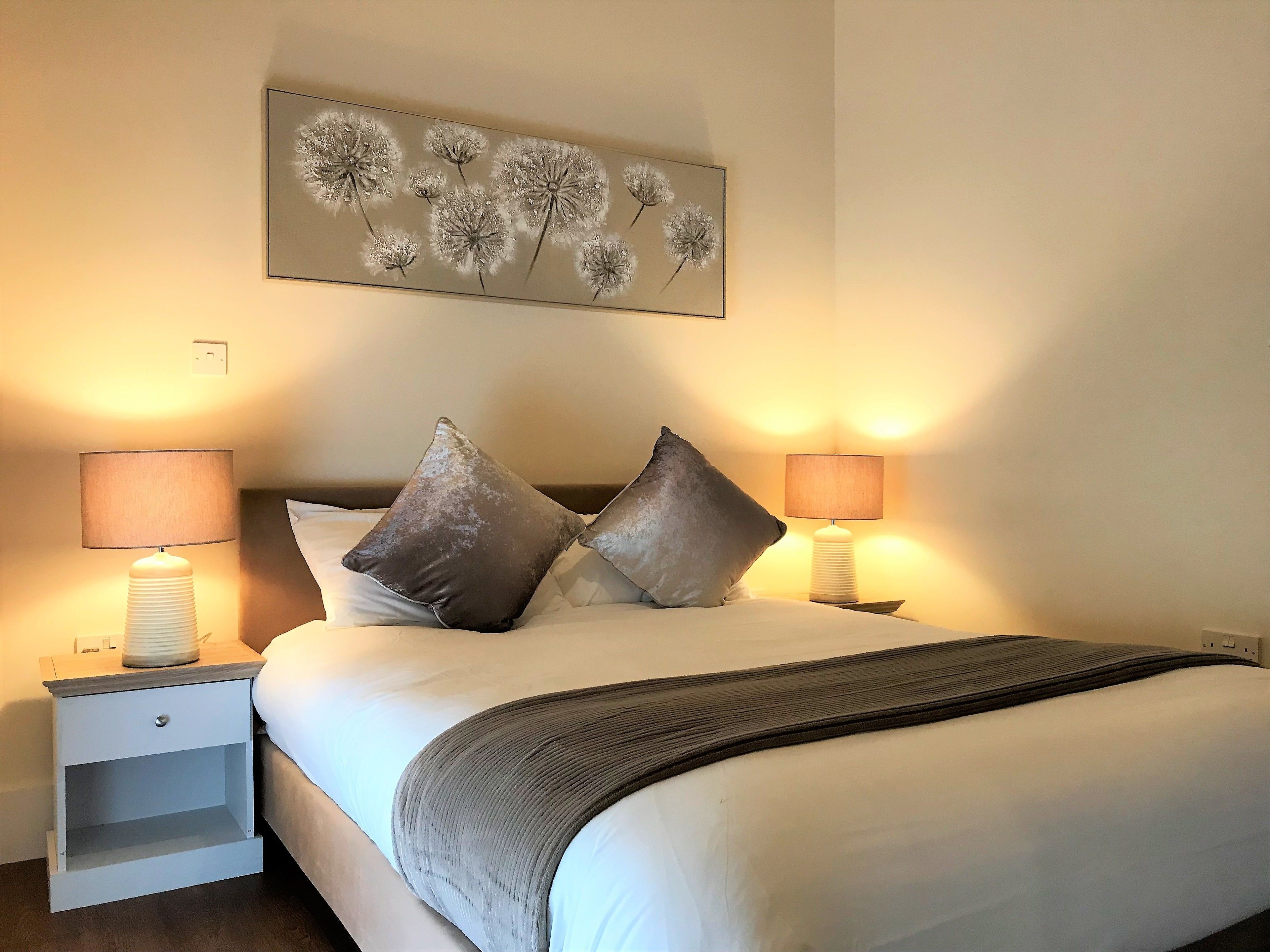 Een net opgemaakt bed met twee kussens, een nachtkastje met een lamp en een wanddecoratie van paardenbloemen.