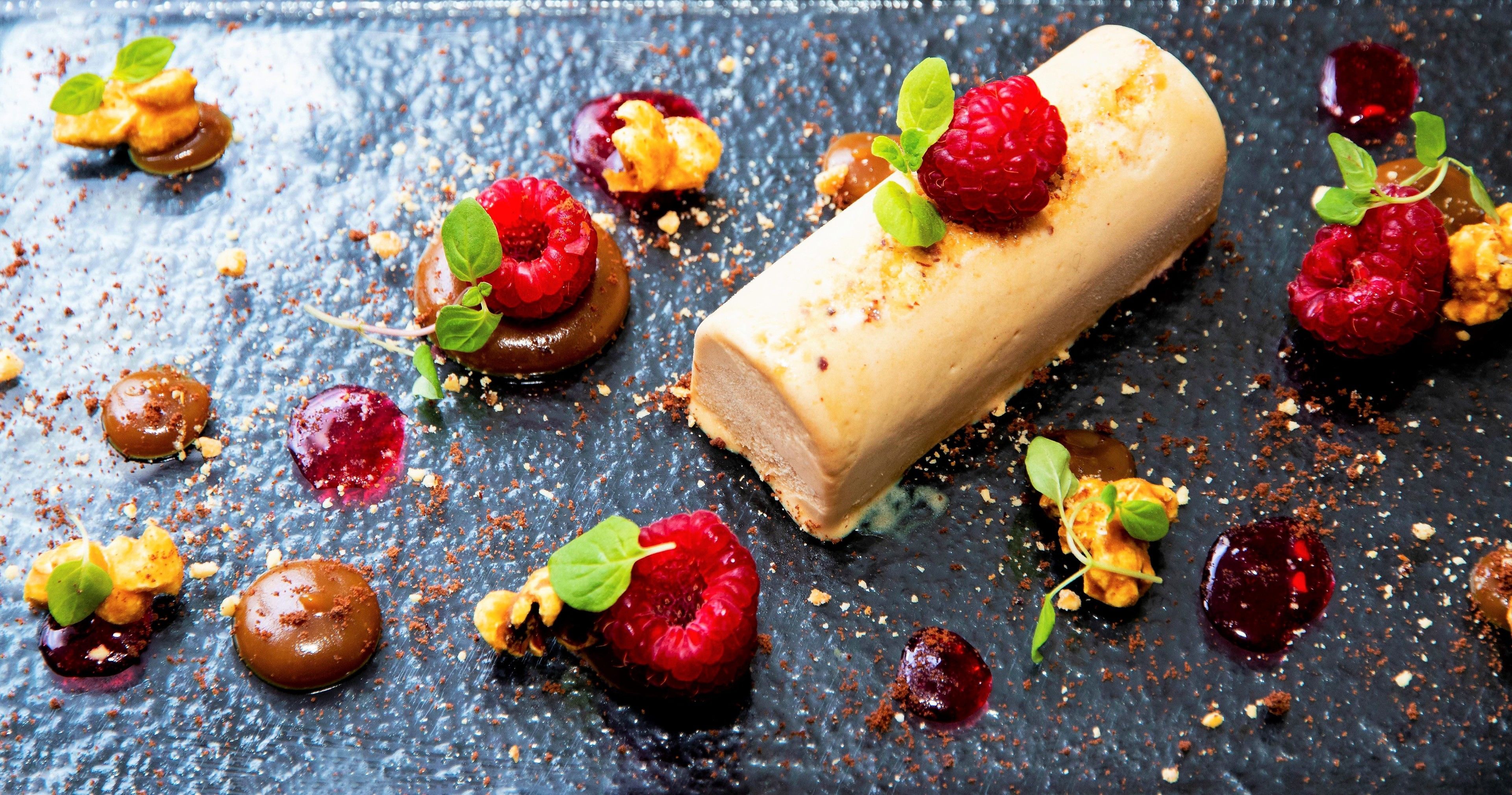 A dessert with a rolled pastry, raspberries, and chocolate sauce on a dark surface.