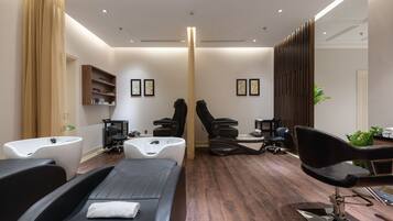 A modern spa room with a massage table, two massage chairs, a small reception desk, and a large mirror.