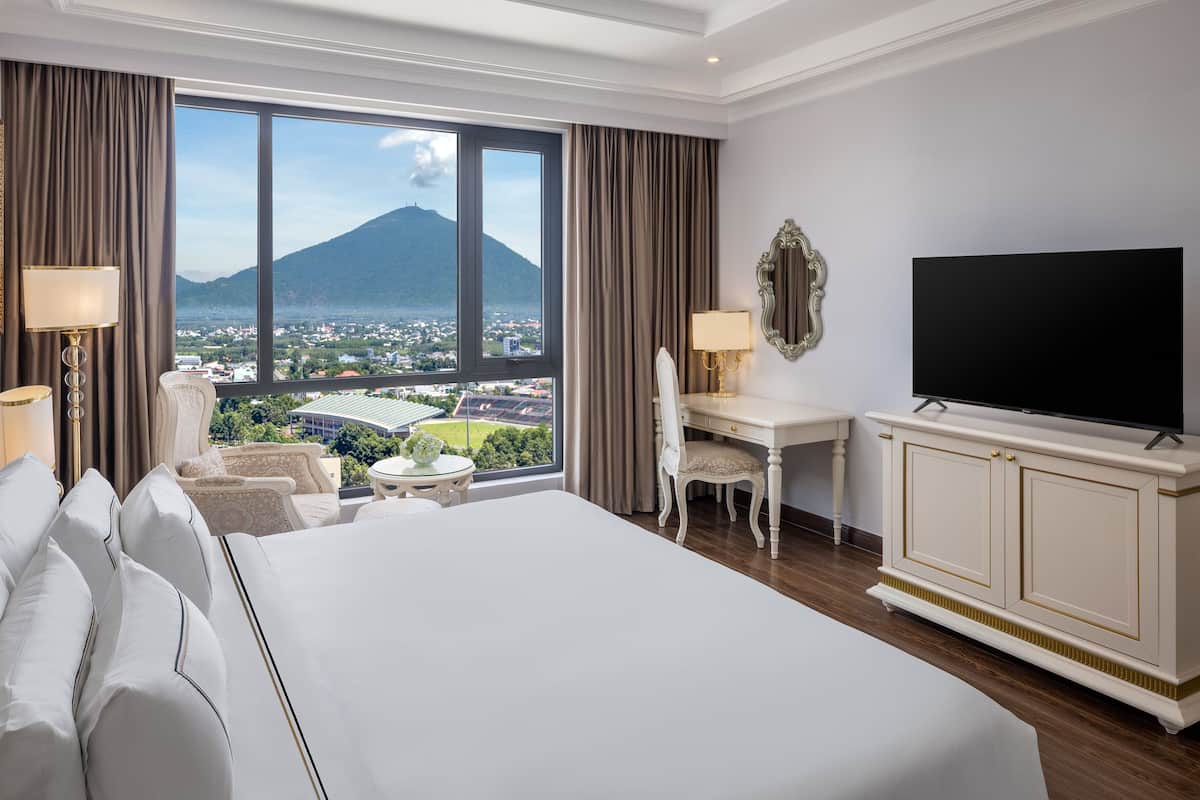 Habitación de hotel con una cama grande, un escritorio, una silla, un televisor y vistas a una montaña y a la ciudad.