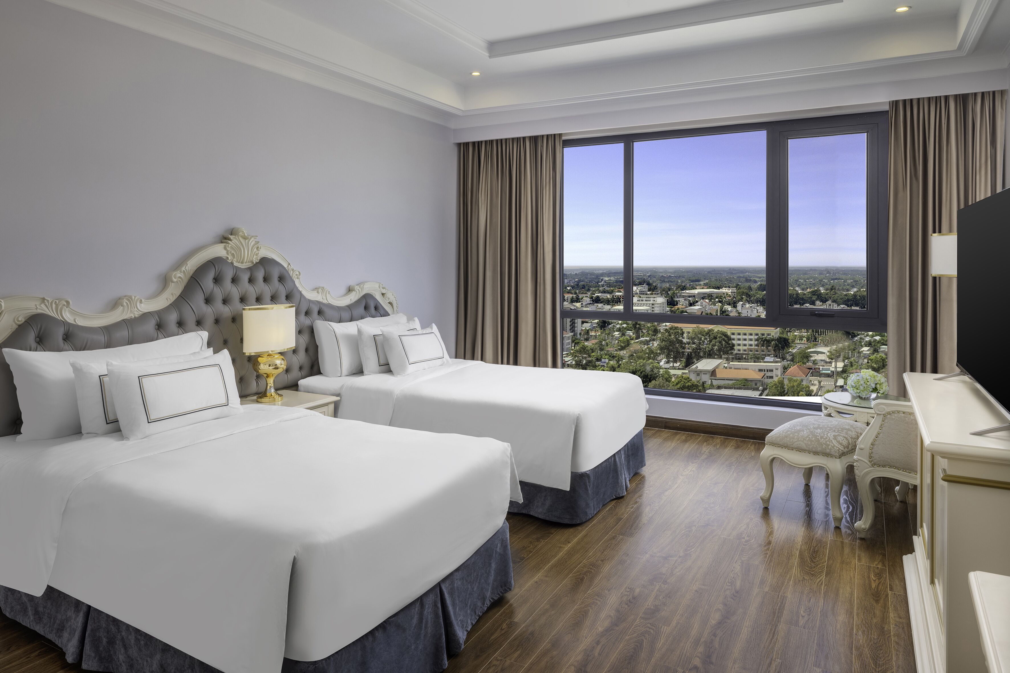 Habitació d'hotel amb dos llits, una finestra gran amb vista a la ciutat, una taula de treball amb una cadira i una televisió.