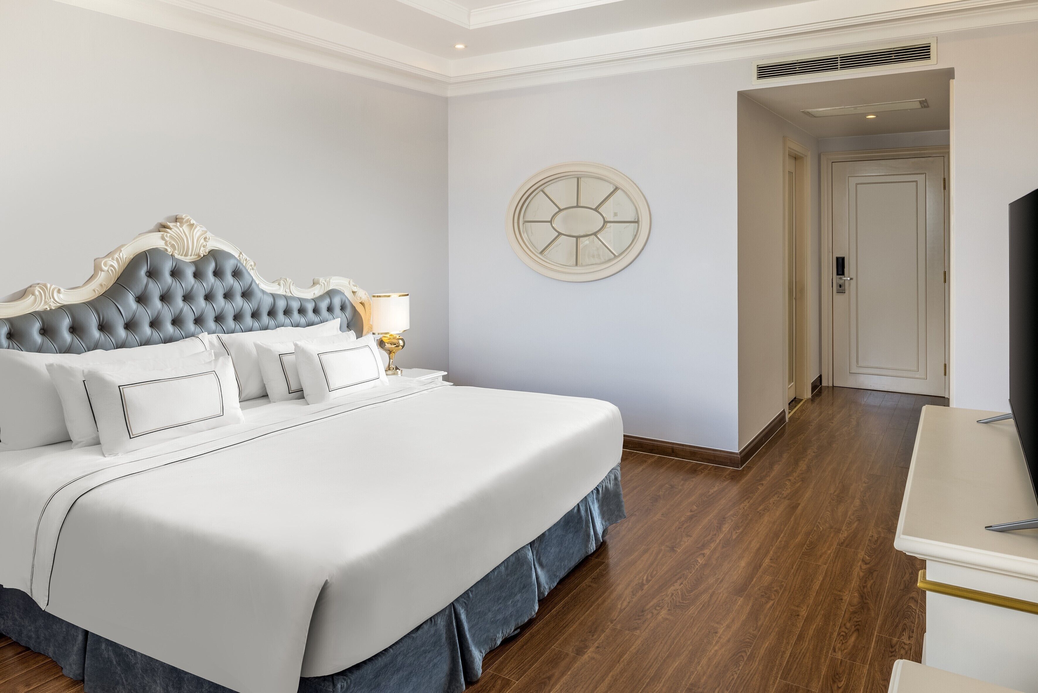 A neatly made bed with a tufted headboard, a bedside table with a lamp, and a round wall clock.