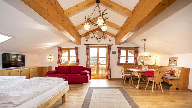 A cozy attic bedroom with a wooden ceiling, a red sofa, a dining table with chairs, and a television on the wall. - Alphirt Ferienwohnungen (Fischen im Allgaeu)