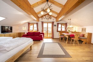A cozy attic bedroom with a wooden ceiling, a red sofa, a dining table with chairs, and a television on the wall.