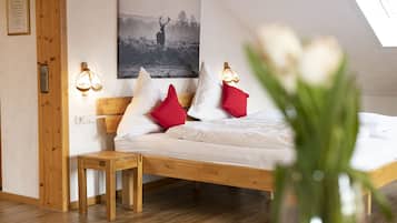 A bedroom with a wooden bed, white bedding, red and white pillows, a small wooden nightstand, a wall-mounted light fixture, and a framed picture above the bed.