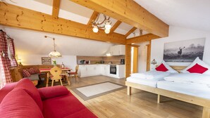 A cozy room with a red sofa, a wooden ceiling, a dining area with a table and chairs, and a kitchen with white cabinets and a stove. - Alphirt Ferienwohnungen (Fischen im Allgaeu)