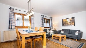 A modern living room with a wooden dining table, a grey sofa, and a wooden coffee table. There are two windows with curtains, a radiator, and a framed picture on the wall. - Alphirt Ferienwohnungen (Fischen im Allgaeu)
