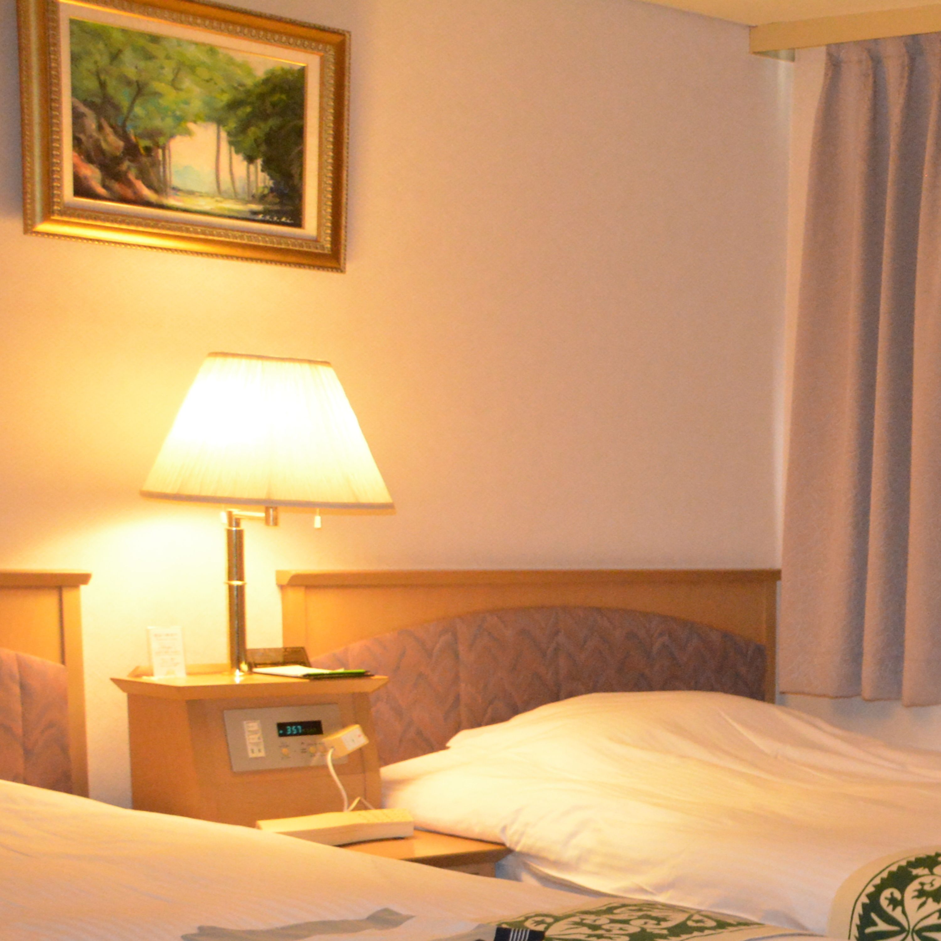 Una habitación de hotel con una cama, una mesita de noche con lámpara, un cuadro en la pared y una ventana con cortinas.