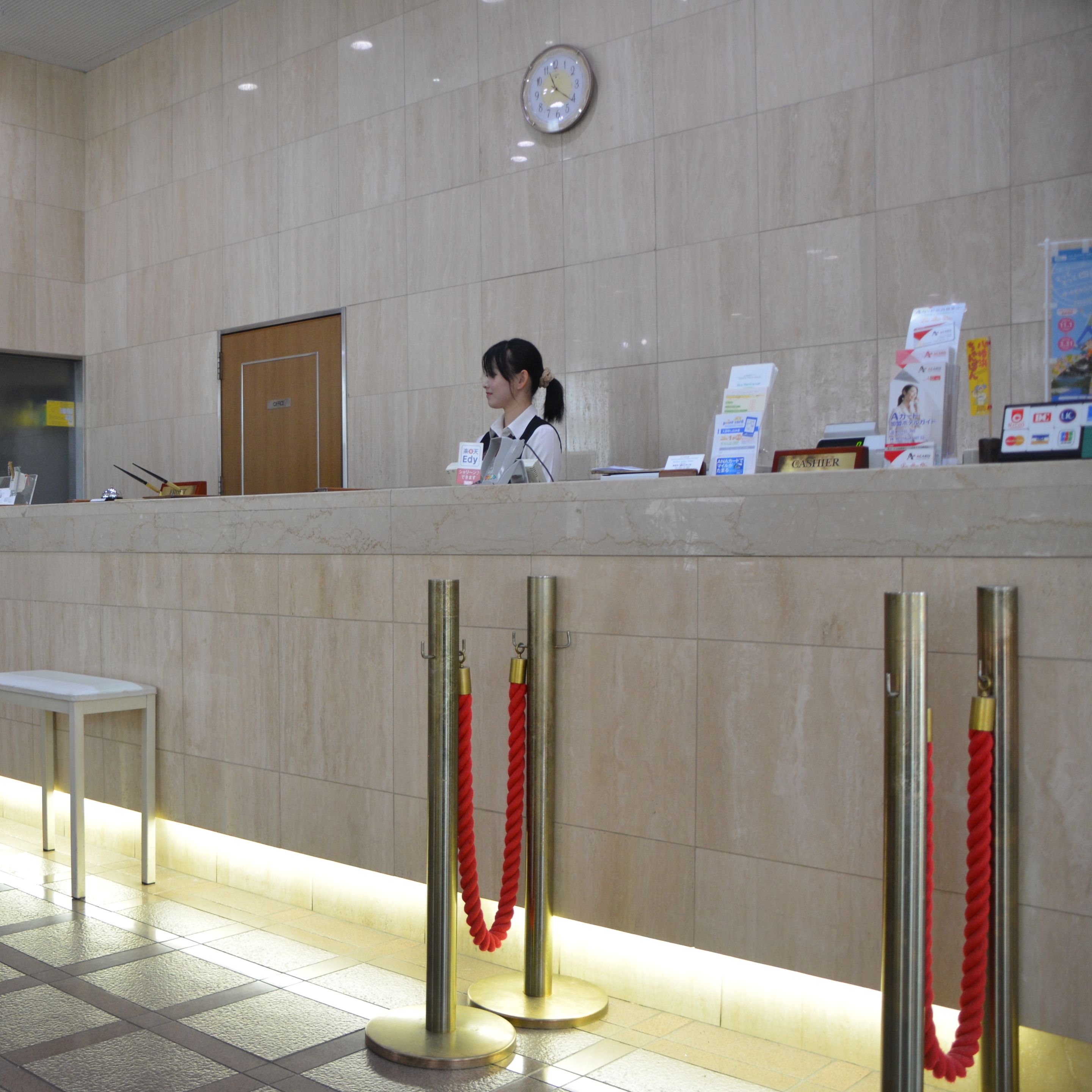 A reception desk with a clock, a woman standing behind it, and red ropes on the floor.