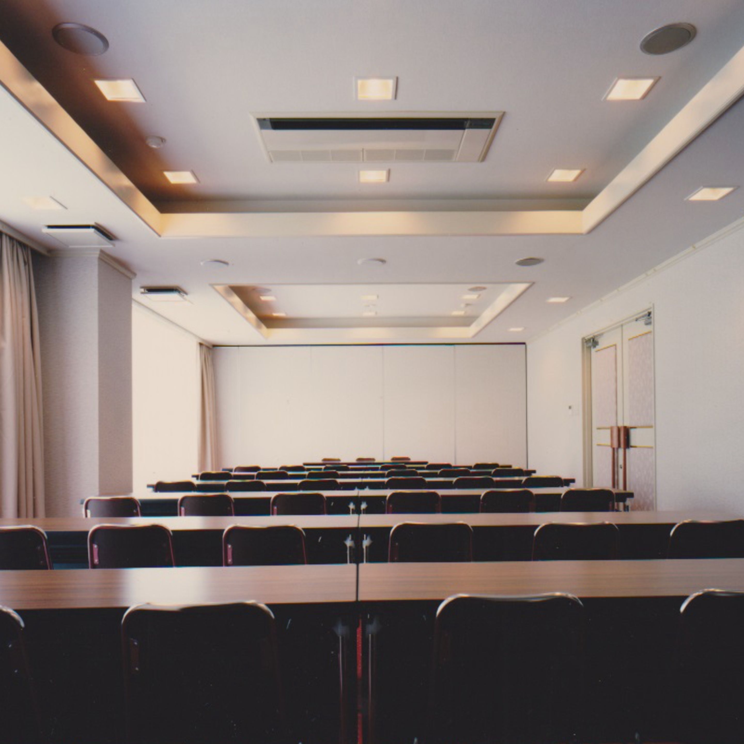 Uma sala de conferências com fileiras de cadeiras, uma mesa comprida e iluminação embutida no teto.