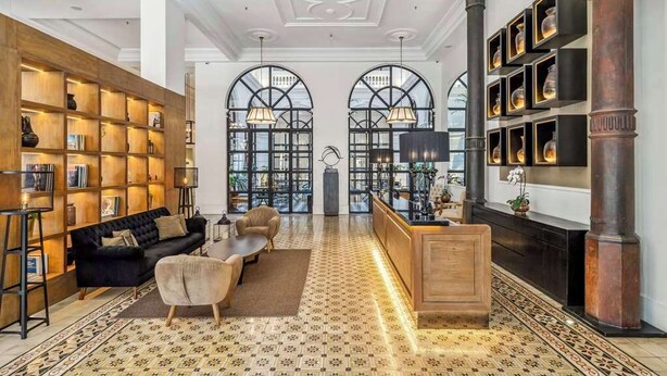 Un vestíbulo amplio con un sofá negro, estanterías de madera, suelo estampado y una mesa de recepción con una gran columna.