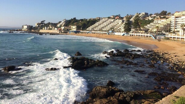 A coastal beach with rocky shoreline, waves crashing onto the shore, and a row of buildings in the background. - Departamento Mares de Concon (Concon)