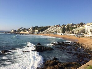 A coastal beach with rocky shoreline, waves crashing onto the shore, and a row of buildings in the background. - Departamento Mares de Concon (Concon)