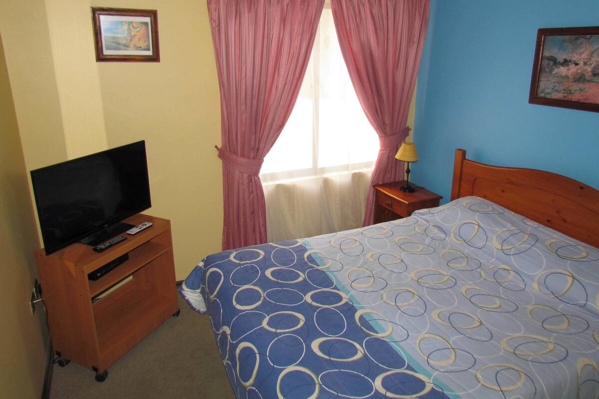 Een hotelkamer met een bed, een televisie op een houten kast, een raam met roze gordijnen en een lamp op een nachtkastje.