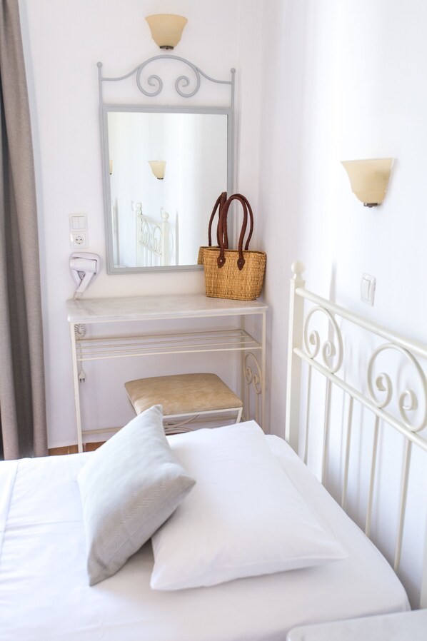 Una cama bien hecha con sábanas y almohadas blancas, una mesita de noche con una cesta y un secador de pelo, y un espejo fijado a la pared sobre un aparador blanco.