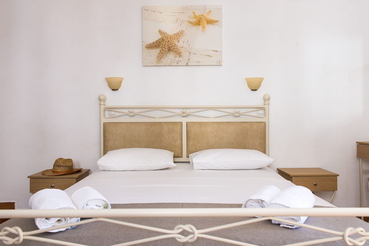 Ein ordentlich bezogenes Doppelbett mit weißen Bettwäsche, zwei Nachttische, ein an der Wand hängendes Kunstwerk mit einem Seestern und ein Strohhut auf einem Tisch.