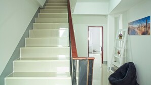 Un escalier avec des marches blanches et une rampe en bois, menant à un palier où se trouvent une étagère blanche et un tableau.