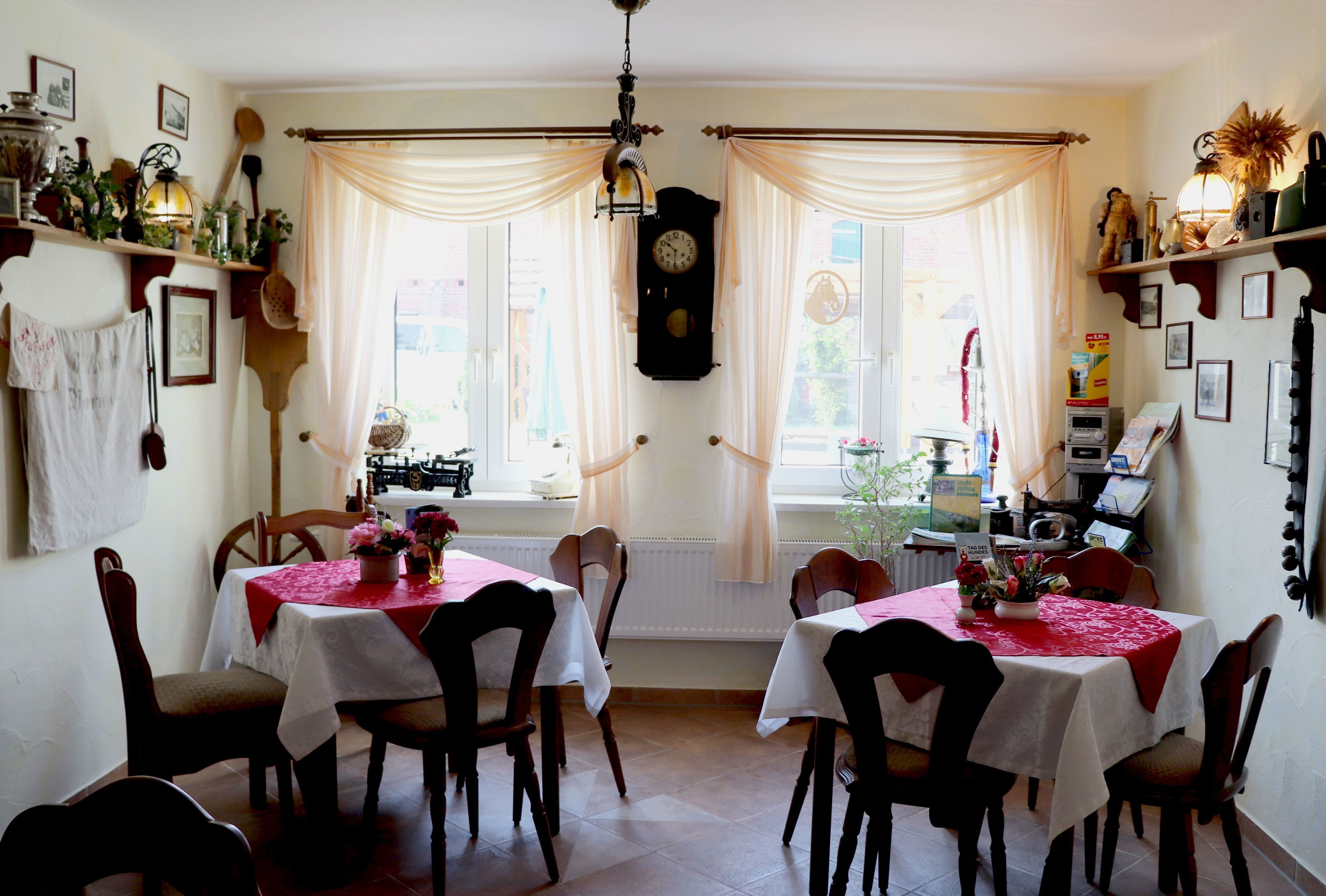A dining room with two tables, each covered with a white tablecloth and red table runners. The room has wooden chairs, a clock on the wall, and a window with sheer curtains.