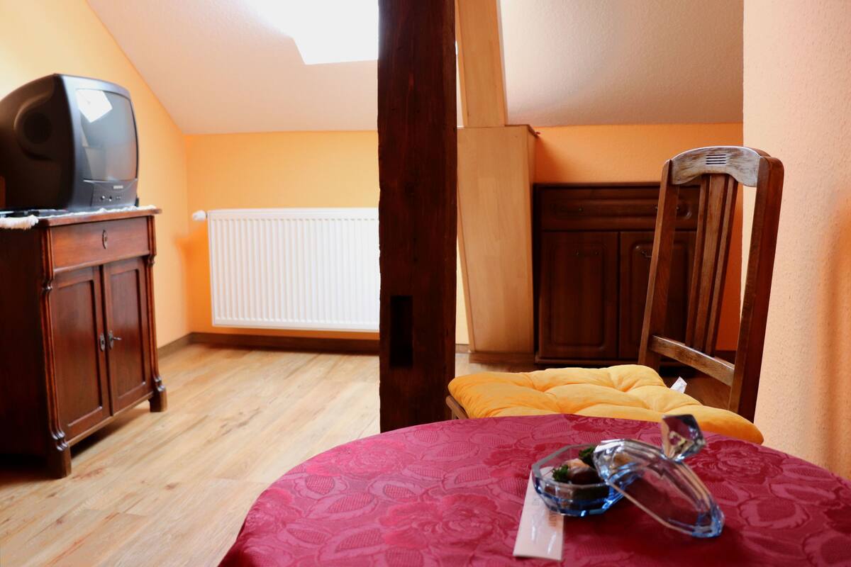 Une pièce avec un meuble en bois, un téléviseur posé dessus, une table rouge sur laquelle se trouve un objet en verre, et un coussin jaune.