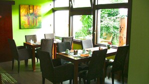 Un comedor con una mesa dispuesta para cuatro personas, rodeada de sillas, ventanas amplias con vista a la vegetación y una pared verde con un cuadro.