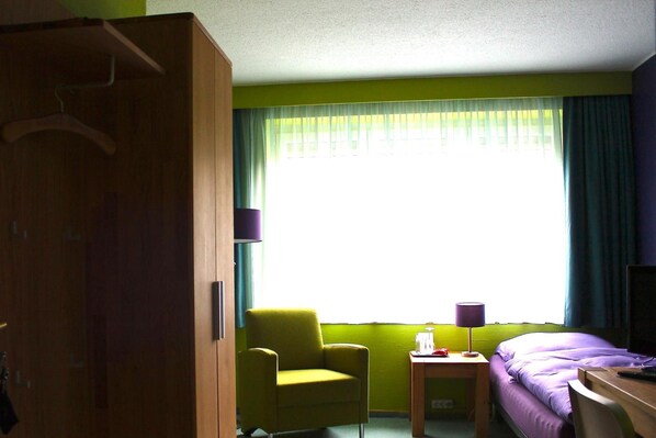 A hotel room with a bed, a chair, a small table, a lamp, and a wardrobe. - Hotel Donnersberg (Darmstadt)