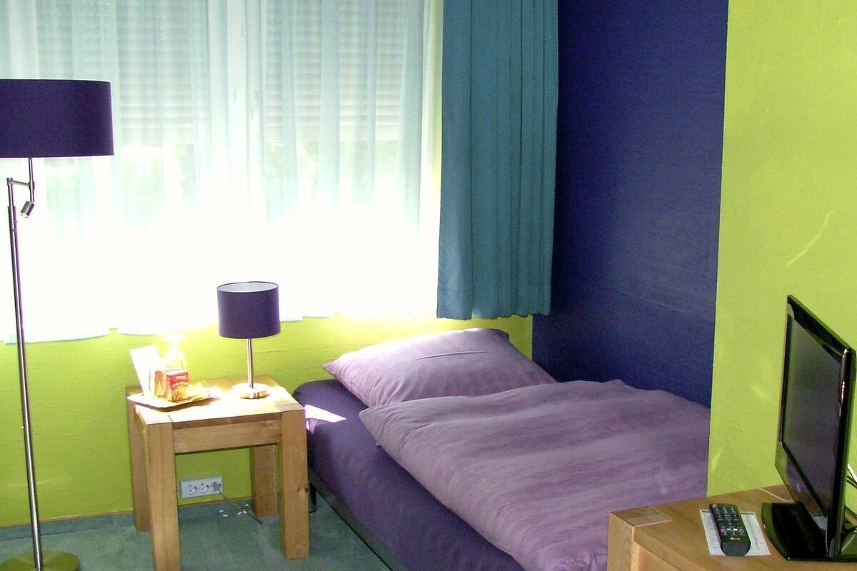 Et lille værelse med en seng, et bord med lampe, et fjernsyn og et vindue med gardiner.