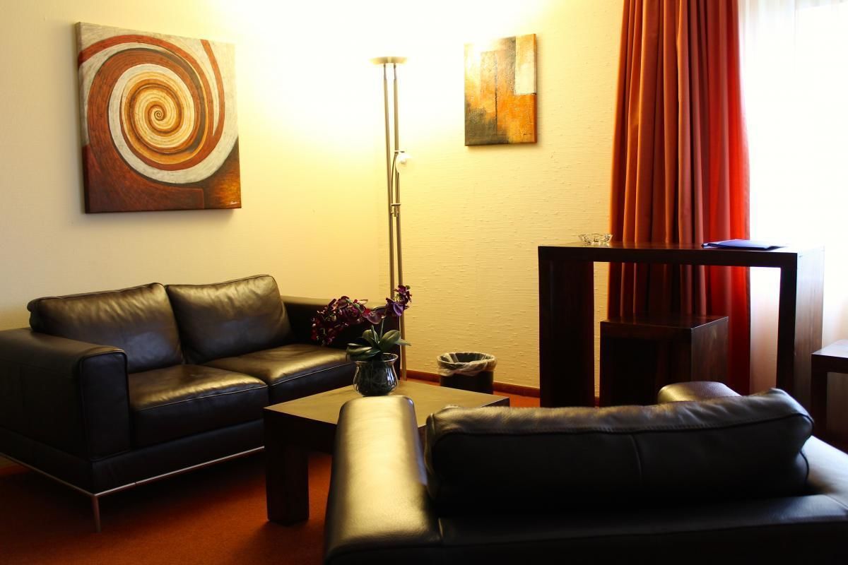 A living room with a black leather sofa, a coffee table, and a floor lamp. There are two paintings on the wall and a red curtain partially drawn.