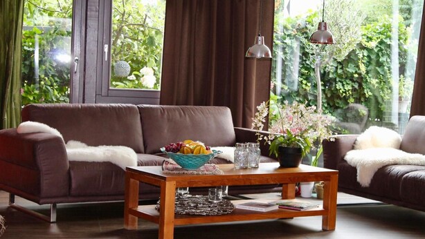 Una sala de estar moderna con un sofá de cuero marrón, una mesa de centro de madera con un frutero, y vistas a zonas verdes a través de amplios ventanales.