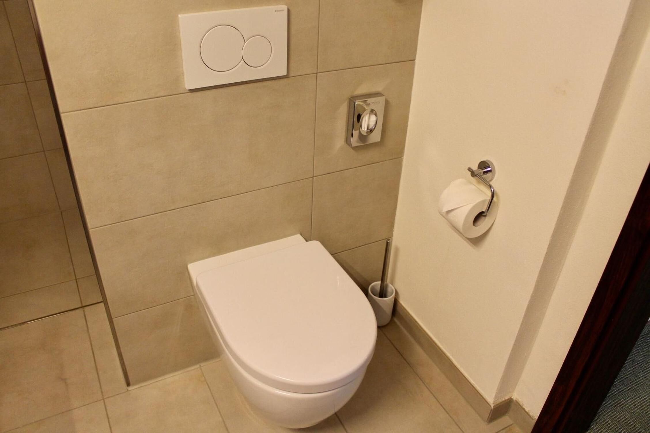 Un baño moderno con un inodoro blanco, un accionador de descarga en la pared y un dispensador de papel higiénico.