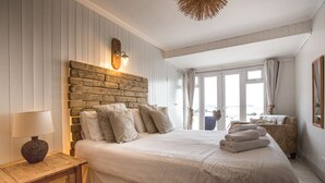 A bedroom with a large bed, wooden headboard, bedside lamp, and a view of the sea through large windows. - Lusty Glaze Beach Accommodation (Newquay)