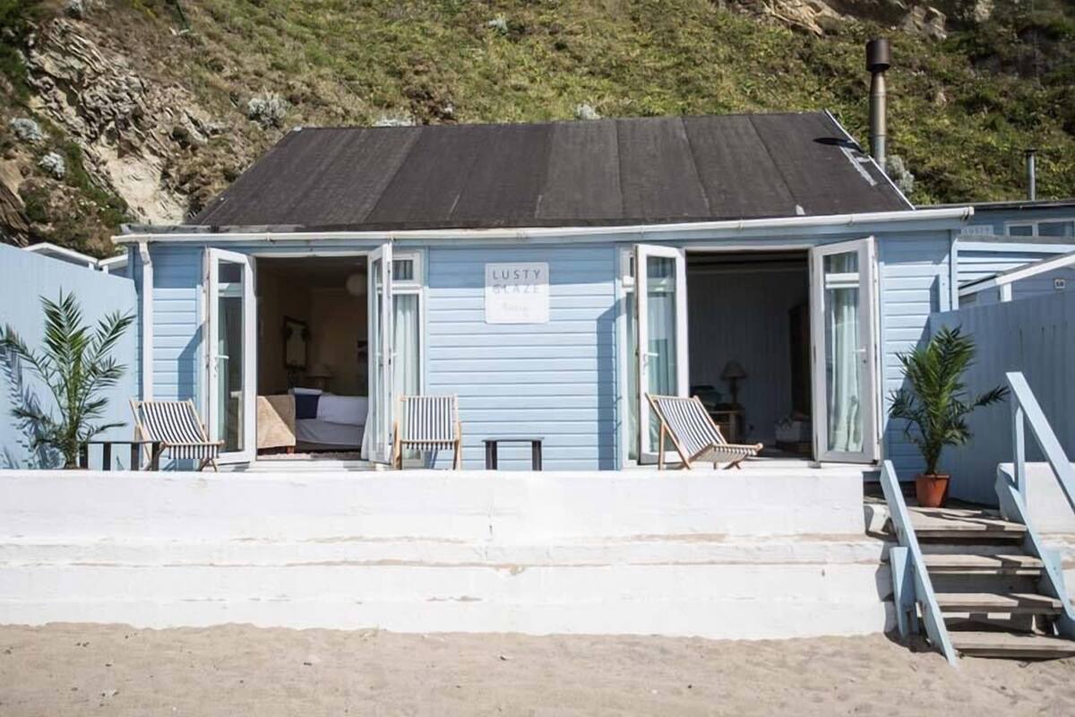 A beach house with an open front door, two chairs outside, and a sign that reads "LUSTY BEACH".
