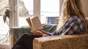 A person reading a book in a room with large windows, a woven chair, and a view of the outdoors. - Lusty Glaze Beach Accommodation (Newquay)