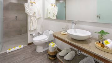 Un baño moderno con inodoro, bidé y un lavabo grande. Hay batas blancas colgadas en un perchero, una zona de ducha y zapatillas en el suelo.