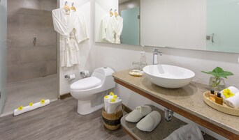 Un baño moderno con inodoro, bidé y un lavabo grande. Hay batas blancas colgadas en un perchero, una zona de ducha y zapatillas en el suelo.
