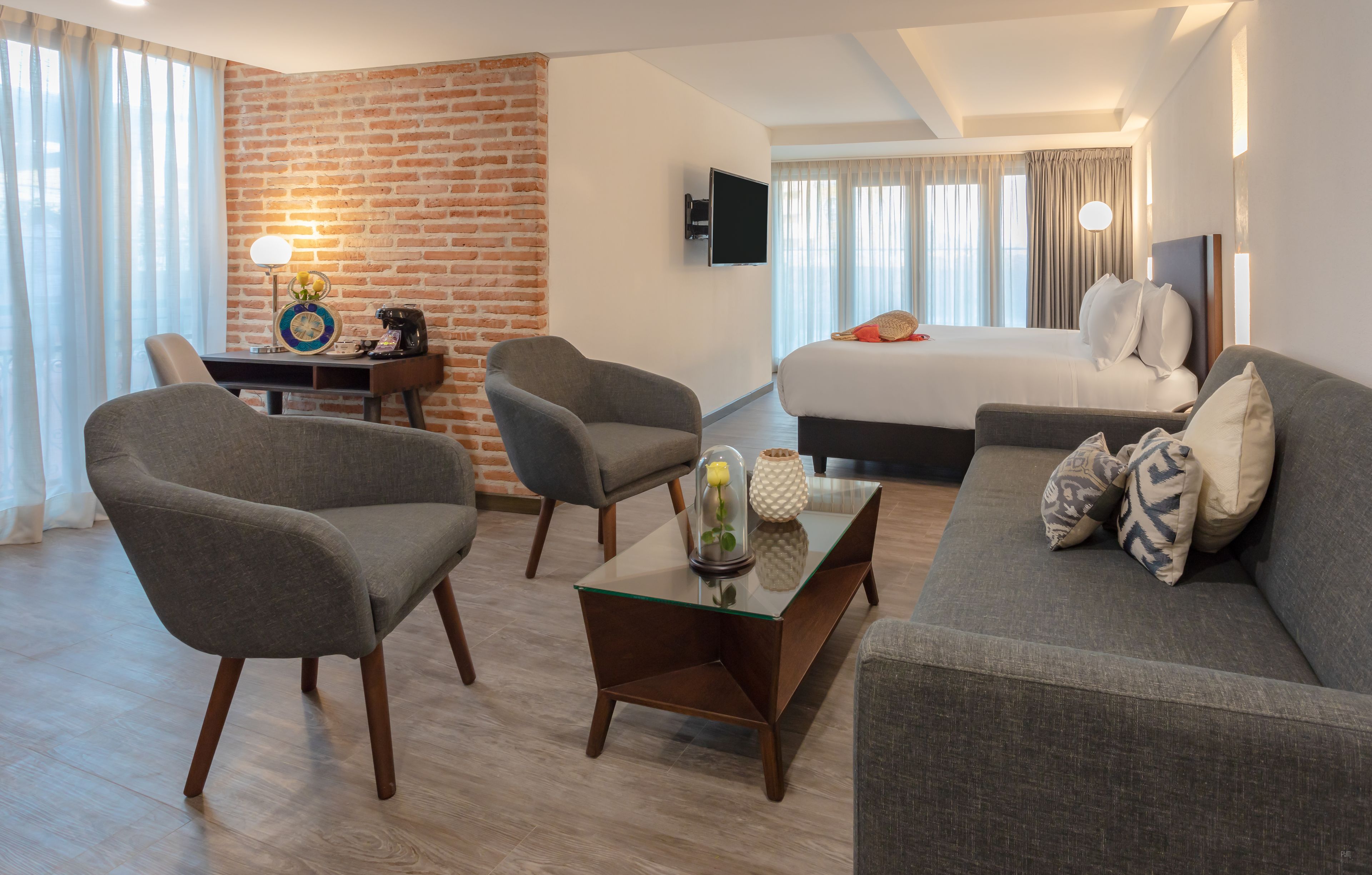 A modern hotel room with a brick accent wall, a large bed, a sofa, two armchairs, a coffee table with decorative items, and a flat-screen TV mounted on the wall.