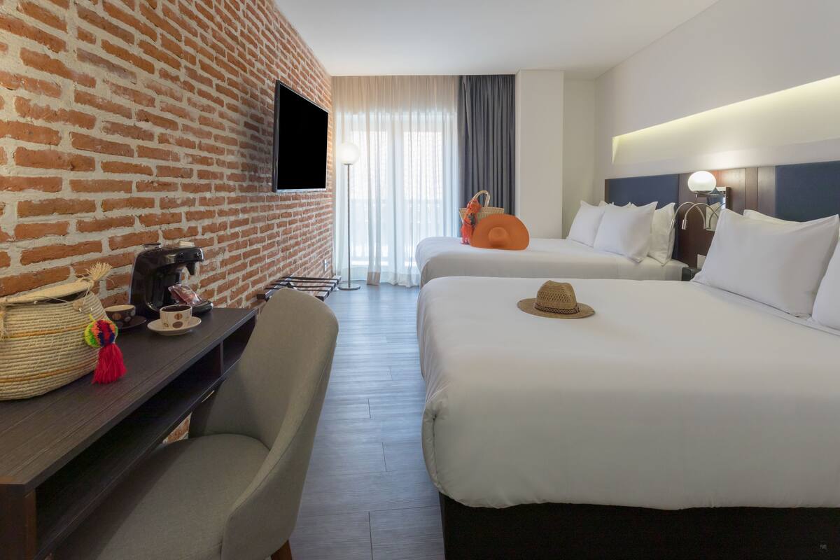 Hotelkamer met twee bedden, een bakstenen muur, een flatscreen televisie, een koffiezetapparaat, een mand en een hoed op het bed.