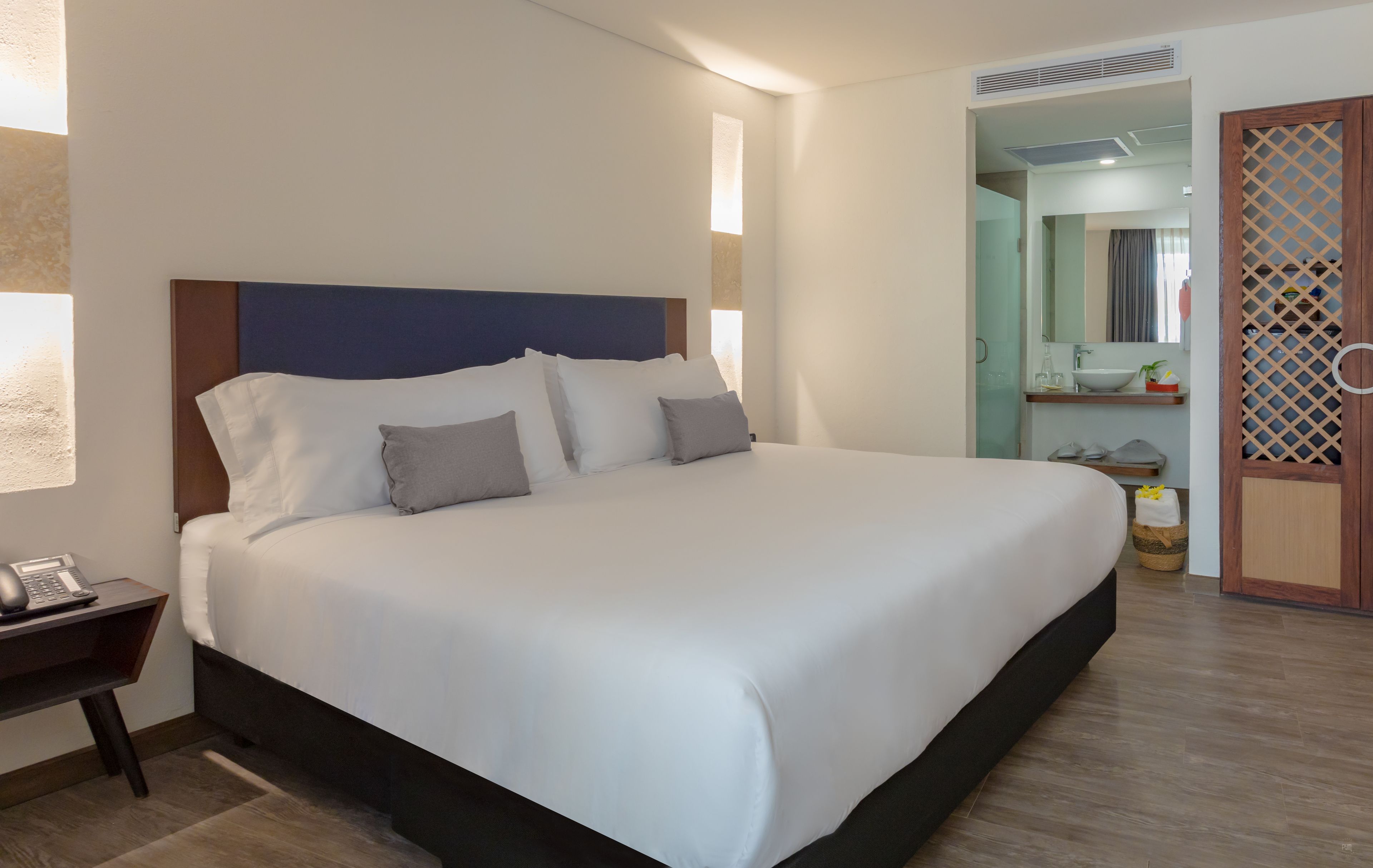 Una cama bien hecha con cabecera de madera, una mesita de noche con teléfono, y un baño visible a través de una puerta abierta.