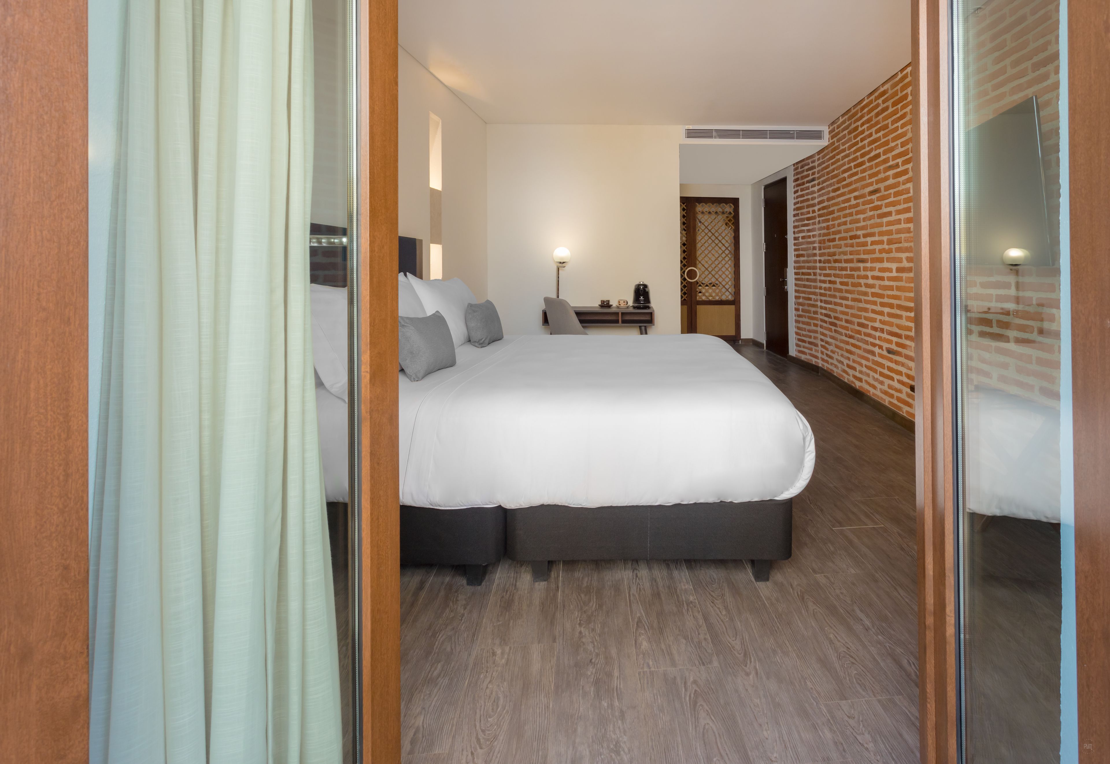 A modern hotel room with a large bed, a desk with a lamp, and a brick wall.