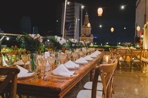 Una larga mesa de comedor dispuesta con copas, servilletas y cubiertos, rodeada de sillas, con vista a un edificio y luces colgantes al fondo.