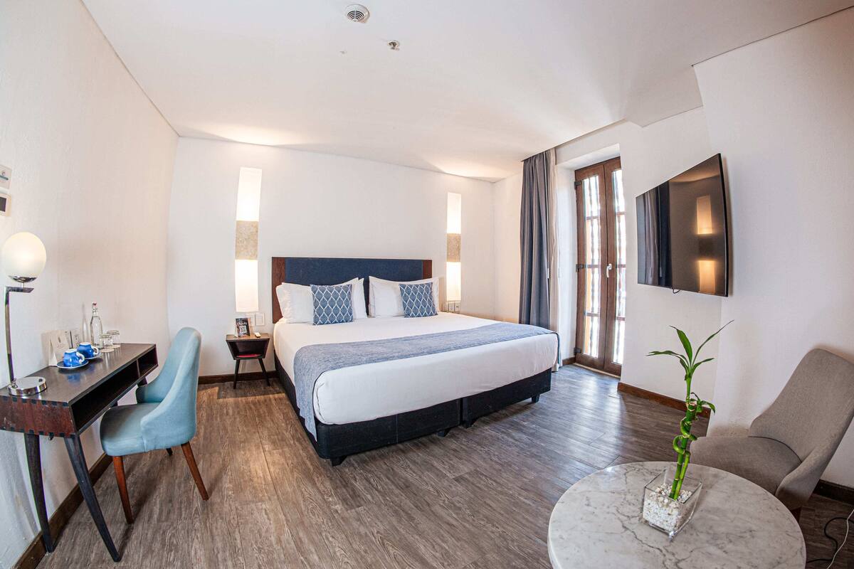 Een hotelkamer met een groot bed, een bureau met stoel, een aan de muur bevestigde televisie en een potplant op een bijzettafeltje.