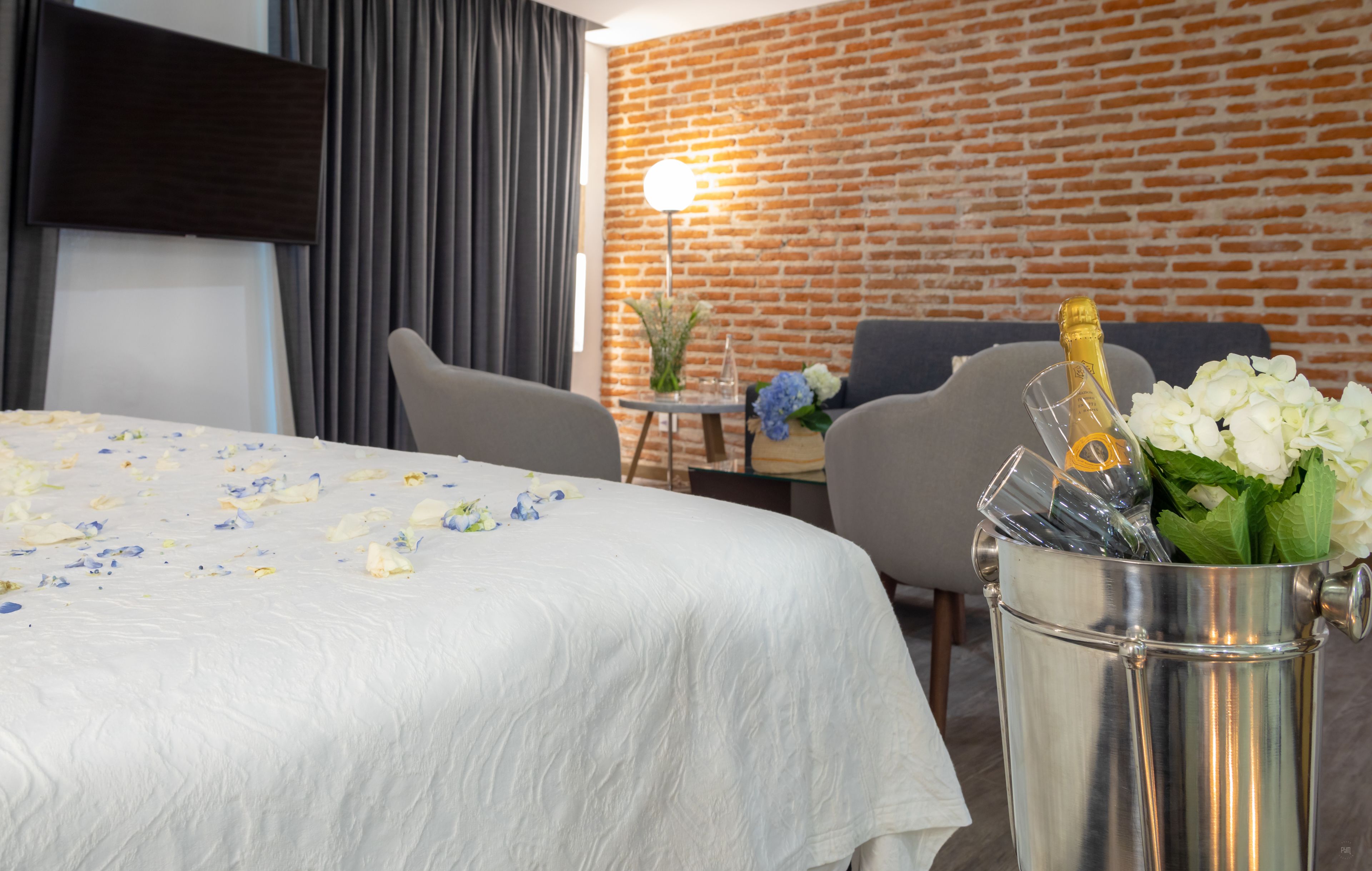 Habitación de hotel con una cama, una mesita con un florero, una silla y un balde con una botella de champán.