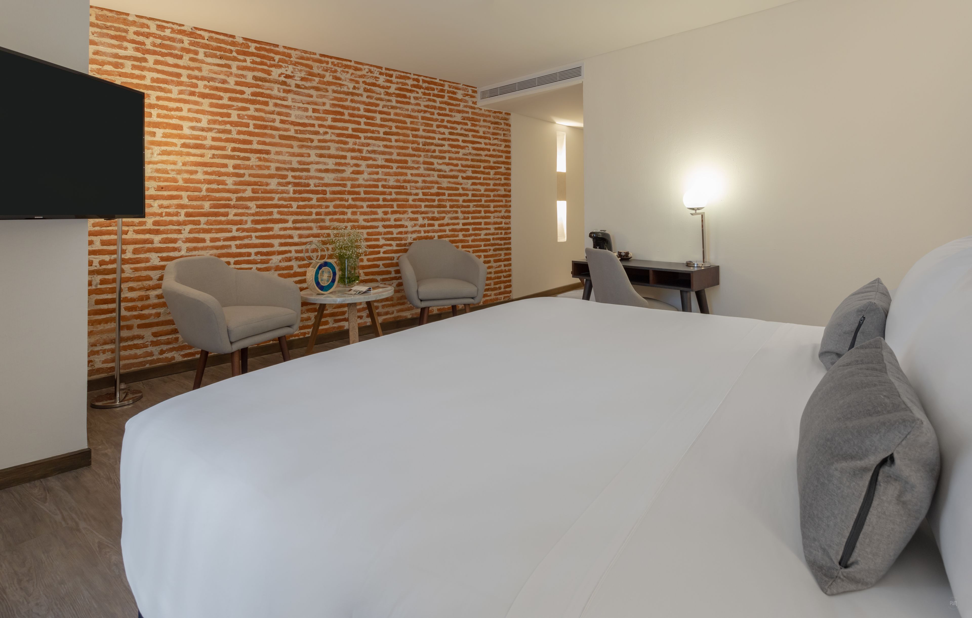 A hotel room with a large bed, two armchairs, a small table with a vase, a desk with a lamp, and a brick-patterned wall.