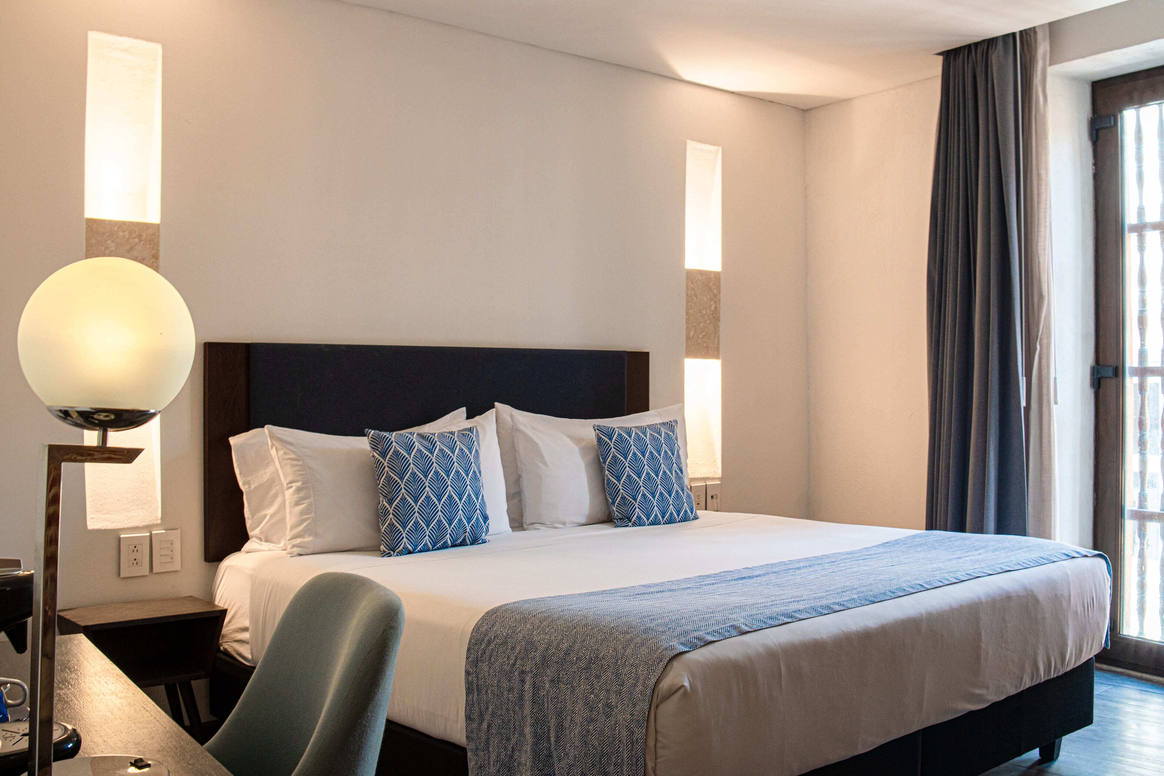A neatly made bed with a dark headboard, white linens, and blue patterned pillows. There is a bedside table with a lamp, and a window with curtains to the right.