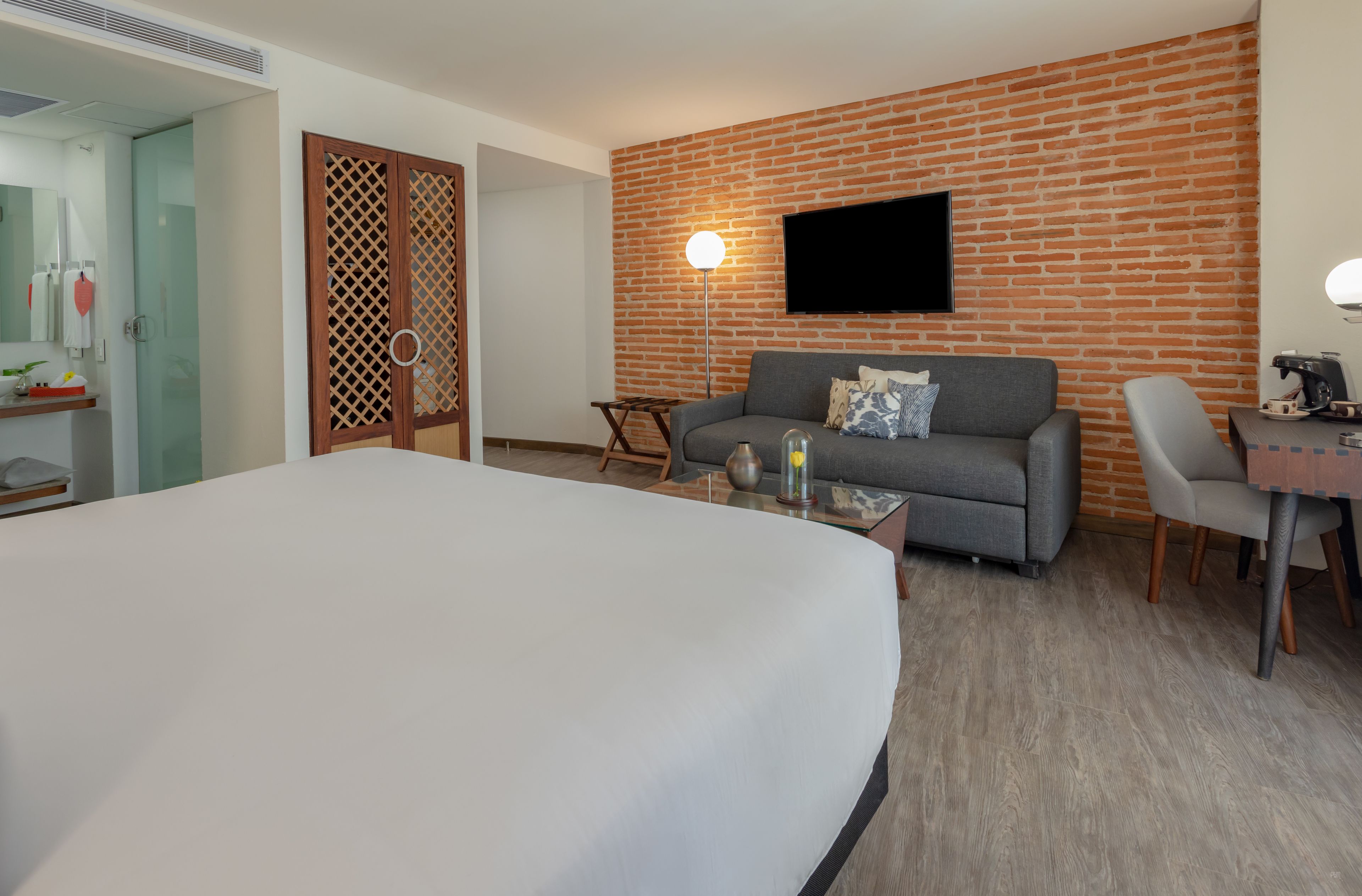 A hotel room with a large bed, a grey sofa, a small table with a lamp, a TV mounted on a brick wall, and a wooden desk with a chair.