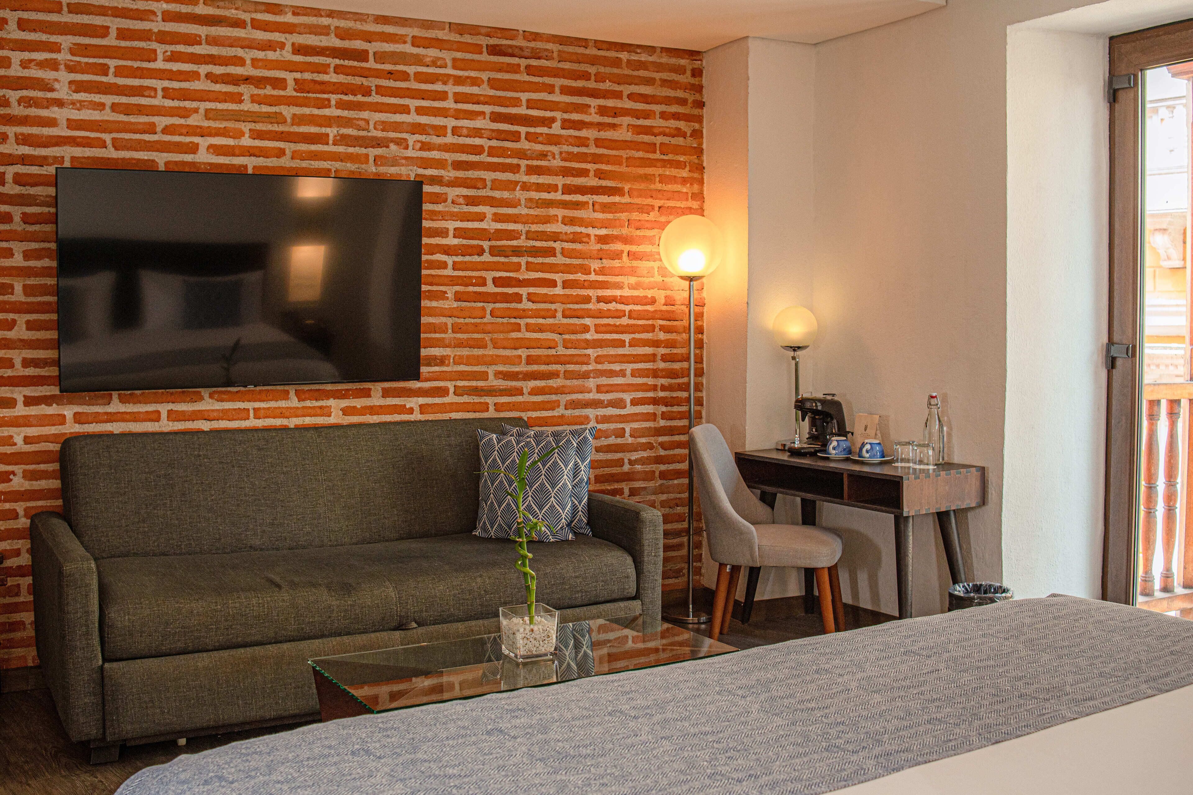 A hotel room with a brick wall, a sofa, a coffee table, a TV, a desk with a chair, and a bed.