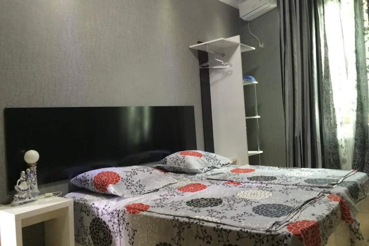 A bedroom with a bed, a nightstand, a wall-mounted shelf, and a window with curtains.